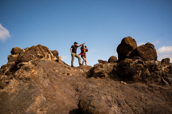 A girl in red shirt with binoculars and a guy pointing her while they are standing on brown desert rocks with blue sky in the backgroungs