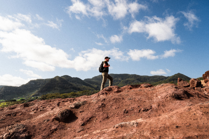 A guy in black t-shirt with a cap and a backpack walks up a rocky hill