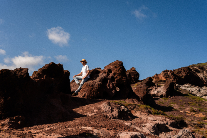 A man in a white shirt with a hat leaning on a sandy rock in a sunny dessert landscape