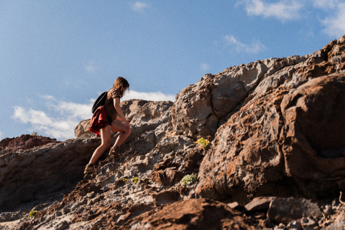 A girl with a backpack climbing up brown desert rocks on a sunny day with blue sky in the background