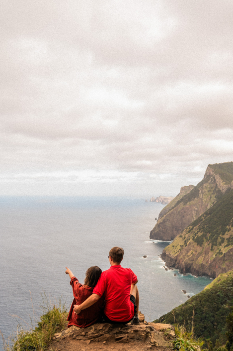 Couple in red shirts sitting on a cliff at the coast line of Madeira island, she is pointing at Porto Santo